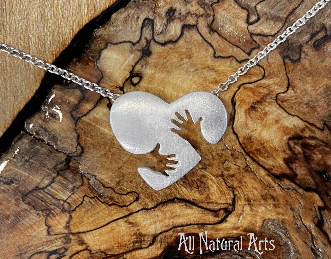 Close-up of the sterling silver Everlasting Hug pendant on silver chain on a wood background, a symbol of love and support, hand-carved and made in New York.