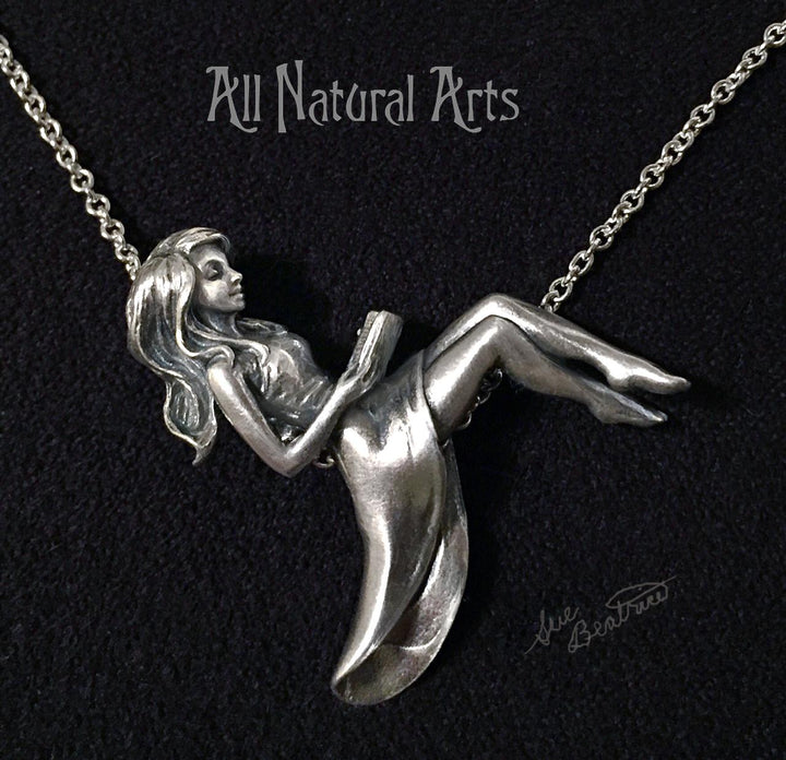Silver pendant of girl reclining on chain reading a book.