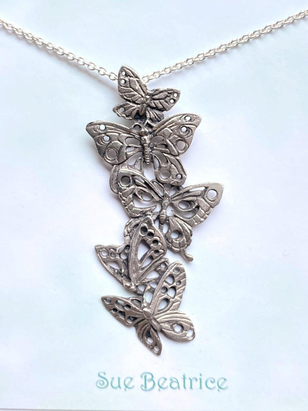A solid sterling silver necklace with five ascending butterflies hanging from the center of a display card. The butterflies are arranged in a filigree design and the necklace is suspended from a sterling silver chain. The display card is square with a light blue cloudy background with Sue Beatrice written underneath.  The item is well-lit and centered on the card.