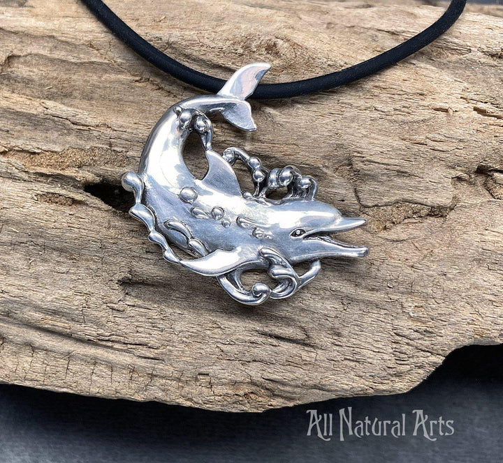 Close-up photo of a sterling, silver dolphin, happily swimming with bubbles coming off his body on a black silicone cord on a driftwood background 