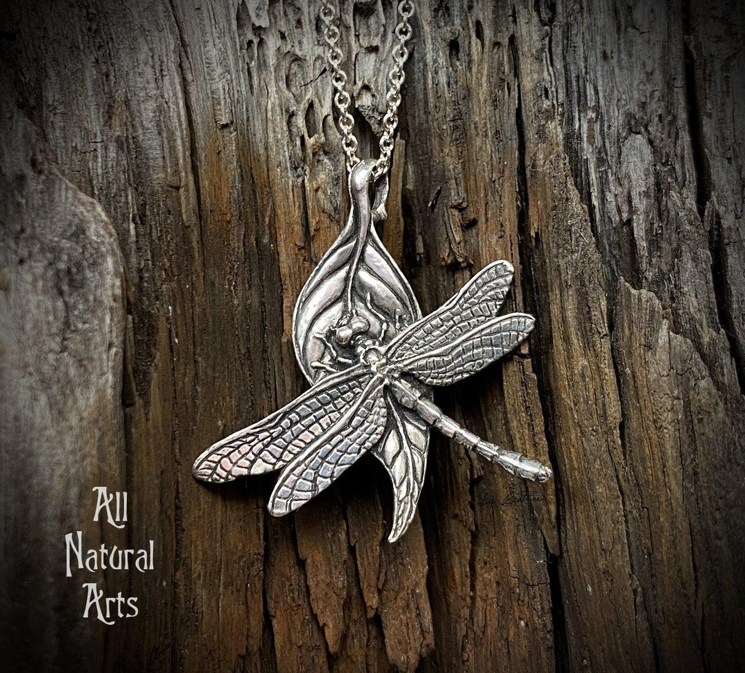 Close-up photo of a beautiful dragonfly, perched on a leaf, which is on a sterling silver chain on a dark wooden background