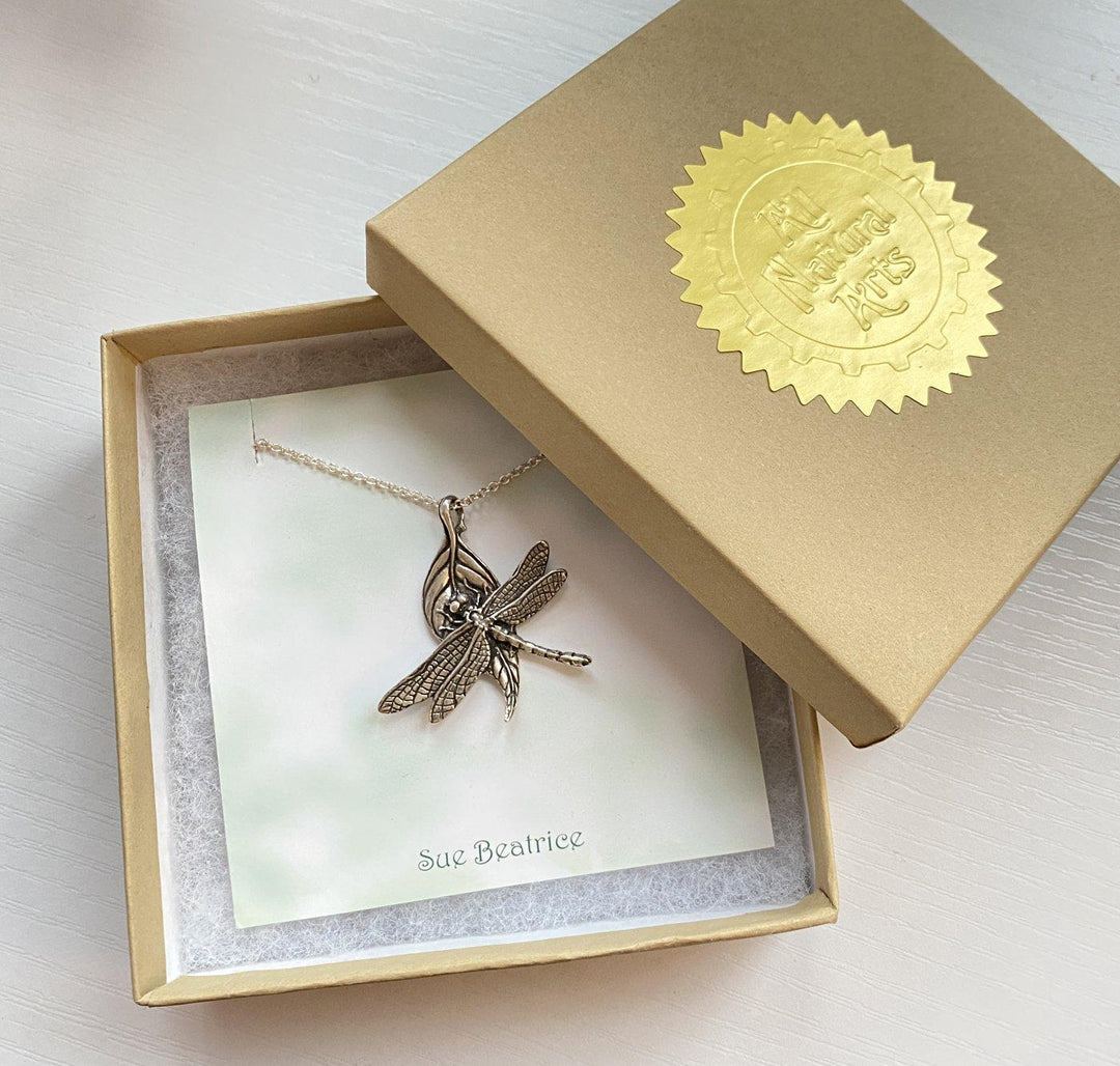 Photo of a open jewelry box with All Natural Arts sticker on front, revealing a beautiful sterling silver dragonfly on a leaf on a sterling silver chain hanging on a card that says Sue Beatrice.  the box is Cotton filled. 