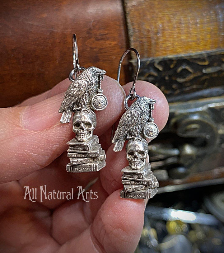 Earrings with raven grasping a pocket watch in its beak, perched atop a skull resting on a stack of books