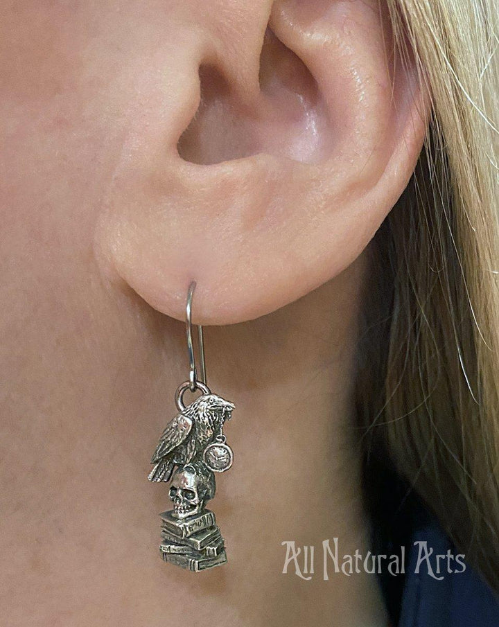 Earrings with raven grasping a pocket watch in its beak, perched atop a skull resting on a stack of books