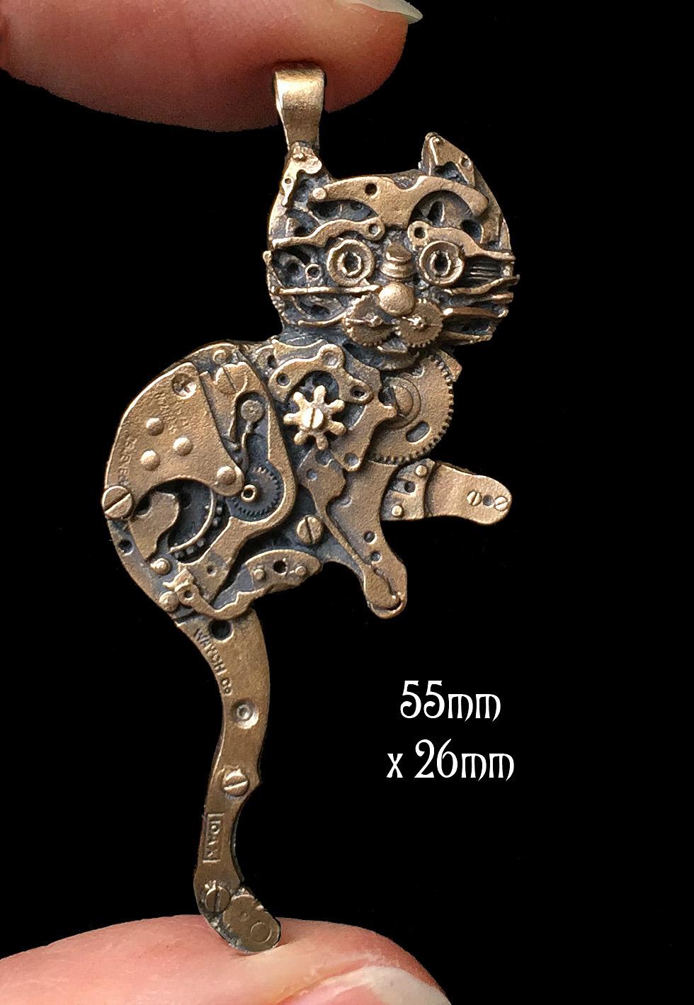 Close up photo of Watch Parts Kitty pendant made of silicon bronze in between fingers, designed by Sue Beatrice of All Natural Arts, featuring intricate details of antique watch parts.